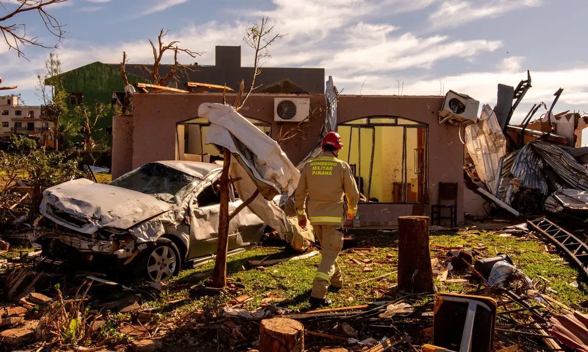 Casa destruída após tornado, com um carro totalmente danificado em primeiro plano e um bombeiro analisando os destroços.